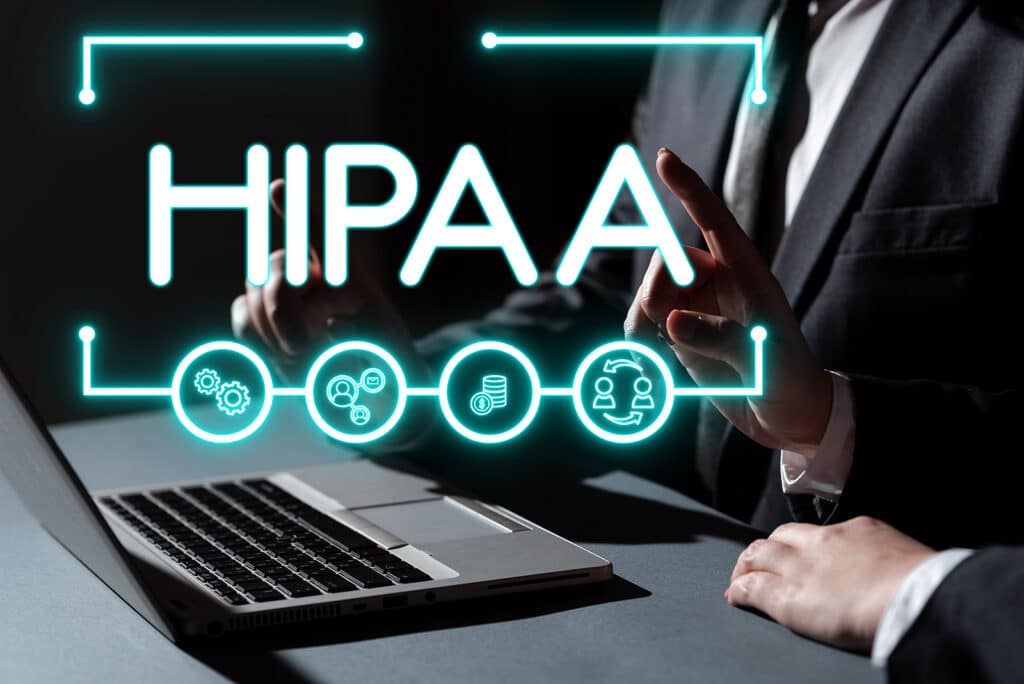 Graphic image of the word HIPAA and various icons over an image of two people's hands at a desk working on a laptop