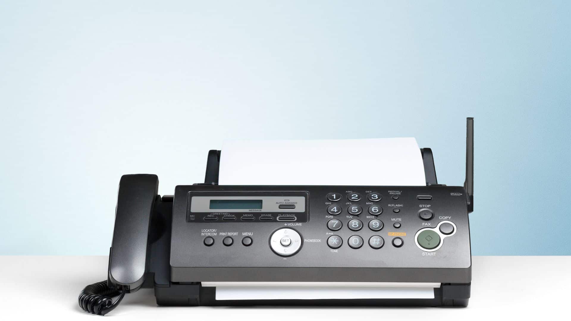 A fax machine with a telephone on a desk, set against a light blue background, used for secure document transmission.