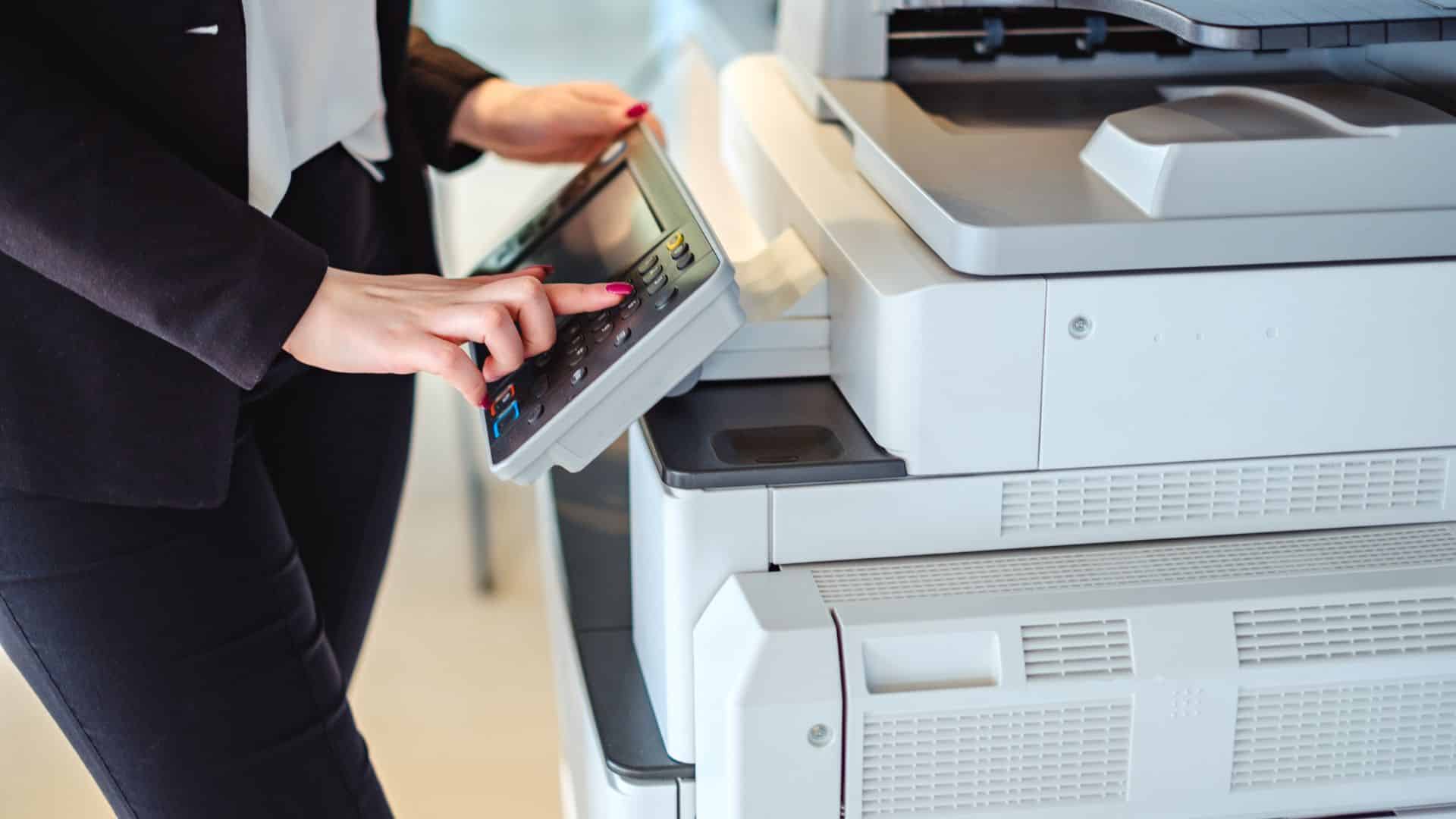 A person using a fax machine in an office, inserting a paper while pressing buttons, with a plant in the background.