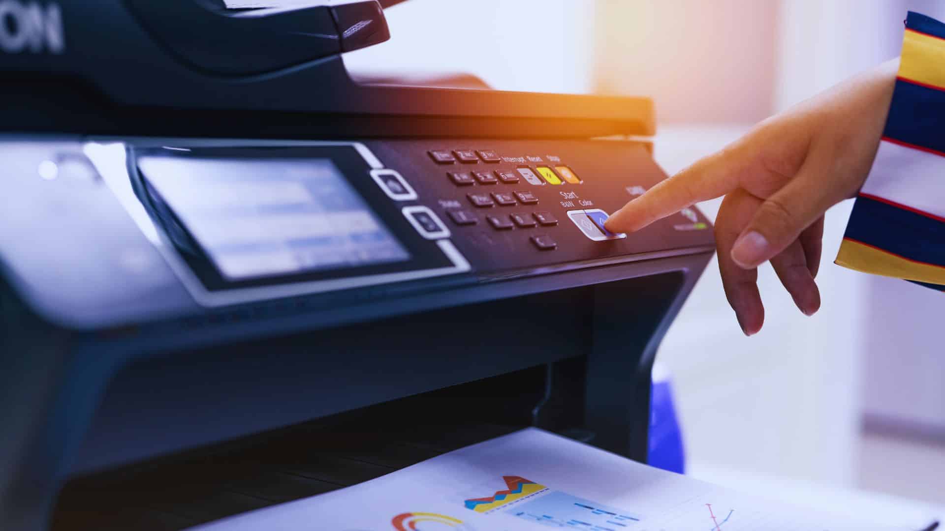 Person pressing the start button on a multifunction printer/scanner to send a fax, with colorful business charts on the paper tray. Healthcare/office fax usage.